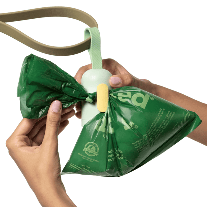 White background with hands holding a dog lead with a light green Earth Rated poop bag dispenser attached. Attached to the dispenser is a poop bag.